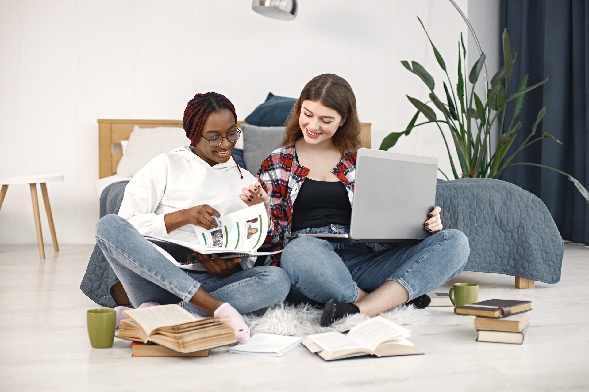Two young teenage girls sitting on the floor near a bed studying and using a laptop